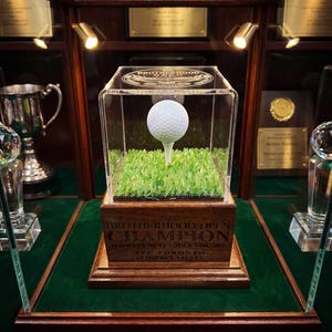 May include: A white golf ball on a tee inside a clear acrylic display case, set on a wooden base with the engraved text: The BROTHERHOOD OPEN CHAMPION (LOWEST NET) - JULY 4TH, 2024 TPC TORONTO AT OSPREY VALLEY. The base features artificial green grass.