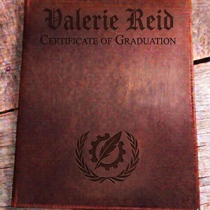 May include: A brown leather certificate holder with the name "Valerie Reid" and "Certificate of Graduation" engraved on the front. A gear and feather emblem is also engraved. The holder is rectangular.