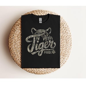May include: A black short-sleeved t-shirt featuring a graphic design of a tiger face outline filled with a camouflage pattern. The text "Tiger PRIDE" is printed in a stylized font with the same camouflage pattern, accompanied by a small paw print.