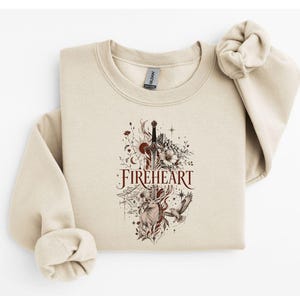 Puede incluir: Una sudadera beige de cuello redondo con un estampado gráfico central que incluye el texto FIREHEART. El diseño presenta una espada, un ciervo, un pájaro, elementos florales, un sol y una luna en tonos rojos, marrones y negros.