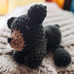 May include: A black and brown crocheted bear plush toy laying on a knitted blanket.