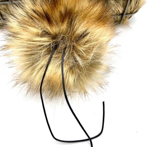 May include: A fluffy brown and blonde faux fur pom pom with black cords attached.