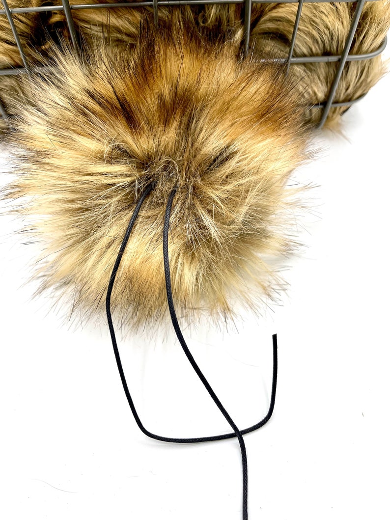 May include: A fluffy brown and blonde faux fur pom pom with black cords attached.