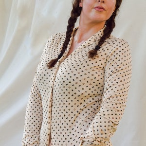 May include: A woman with long brown braids wears a beige button-down shirt with a black and brown geometric pattern. The shirt has long sleeves and a collared neckline.
