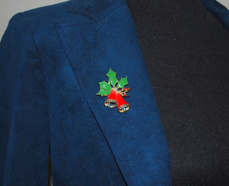 May include: A gold-tone Christmas brooch with red bells and green holly leaves. The brooch is adorned with sparkling rhinestones.