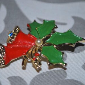 May include: A gold-tone Christmas pin with red bells and green holly leaves. The bells have blue rhinestones and a white pearl accent.