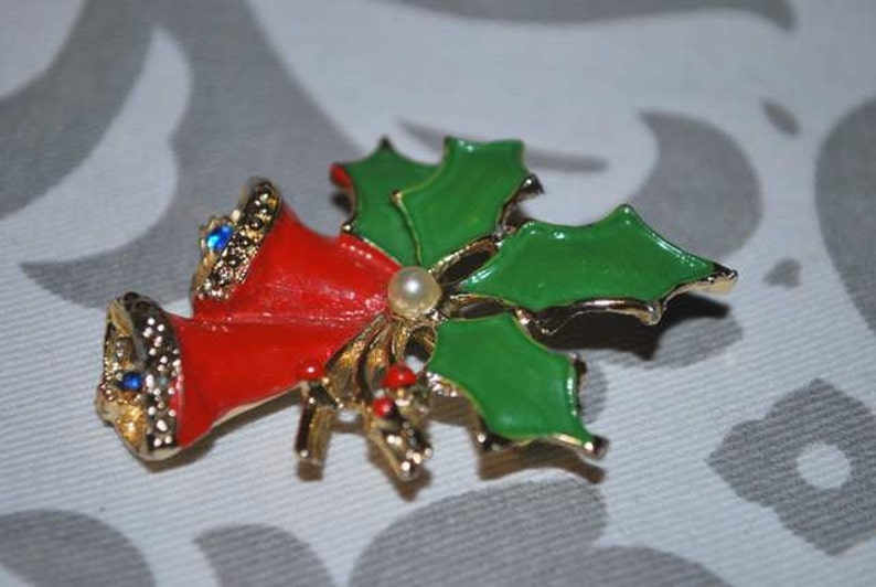 May include: A gold-tone Christmas pin with red bells and green holly leaves. The bells have blue rhinestones and a white pearl accent.