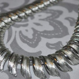 May include: A silver tone necklace with a unique design of overlapping, textured, curved pieces. The necklace is made of metal and has a shiny finish.