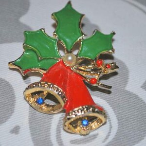 May include: A gold-tone Christmas brooch featuring two red bells with blue rhinestones, surrounded by green holly leaves and red berries.