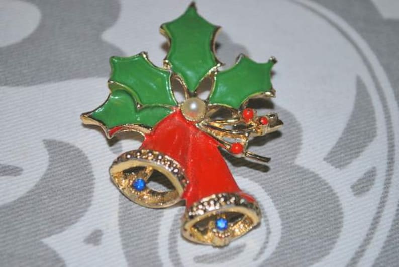 May include: A gold-tone Christmas brooch featuring two red bells with blue rhinestones, surrounded by green holly leaves and red berries.