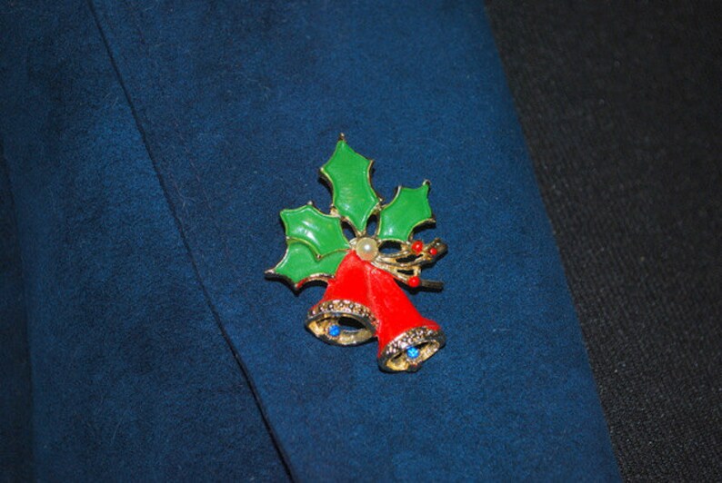 May include: A gold-tone Christmas pin with red bells and green holly leaves. The bells have blue rhinestones and a white pearl accent.