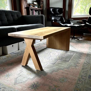 May include: A light wood coffee table with an X-shaped base. The table has a natural wood finish and is sitting on a patterned rug in a living room.