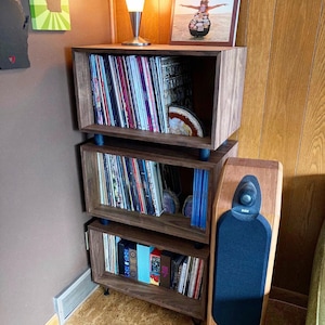 May include: A three-tiered wooden shelf unit with a dark brown finish. The shelves are filled with vinyl records and other items. A speaker is on the right side of the shelf unit.