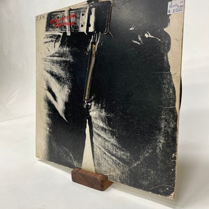 May include: A black and white vinyl record album cover featuring a close-up of a person wearing jeans with a zipper. The album cover is titled "The Rolling Stones - Sticky Fingers" and has a price tag that says "Plays Fine $20.00".