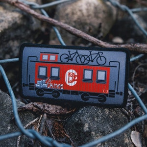 May include: A red and gray embroidered patch with a cartoon train car with two bicycles on top. The train car has the text "End of the Line" and "Campout" on the side. The train car has a number "C3" on the side.