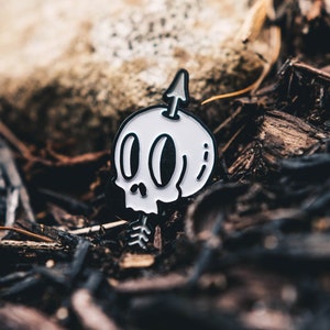 May include: A white enamel pin with a black outline of a skull with an arrow through it. The pin is on a bed of brown wood chips.