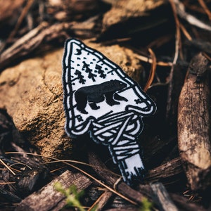 May include: A white and black embroidered patch shaped like an arrowhead with a bear silhouette inside. The bear is walking through a forest with trees and stars. The patch is on a bed of brown pine needles.