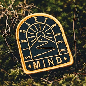 Peut inclure: Un écusson brodé bleu foncé avec une bordure jaune. L'écusson représente une chaîne de montagnes avec un soleil qui se lève au-dessus. Le texte "SINGLETRACK MIND" est écrit autour de la bordure.