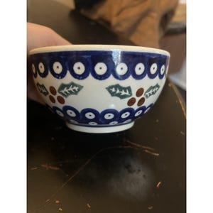 May include: A small ceramic bowl with a cream-coloured base featuring a hand-painted pattern of dark blue circles with white centres, interspersed with green holly leaves and clusters of three brown berries arranged in a horizontal band.