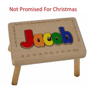 Pode incluir: Banco de madeira com um quebra-cabe&ccedil;a de nome personalizado no topo. O nome "Jacob" &eacute; escrito em letras coloridas. O banco tem quatro pernas e os n&uacute;meros de 1 a 10 est&atilde;o gravados no topo.