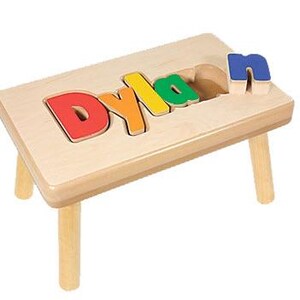 Pode incluir: Um banquinho de madeira com um recorte para o nome "Dylan" em letras coloridas. As letras s&atilde;o remov&iacute;veis e podem ser usadas como um quebra-cabe&ccedil;a.
