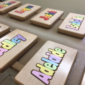 May include: Wooden name puzzle pieces in rainbow colors, spelling out the names "Logan", "Josiah", "Isabella", "Adley", and "Adelaide".