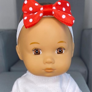 May include: A doll with brown eyes and a red bow with white polka dots. The bow is attached to a white elastic headband. The doll is wearing a white top.