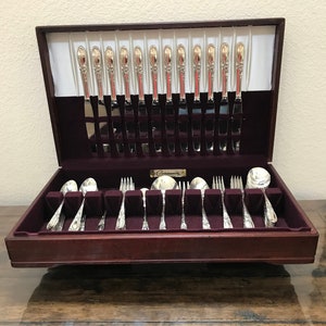 May include: A wooden silverware chest with a burgundy velvet interior, holding a complete set of silver-colored flatware. The set includes knives, forks, spoons, and serving pieces. The chest is open, revealing the neatly arranged utensils. The brand name "Community" is visible.