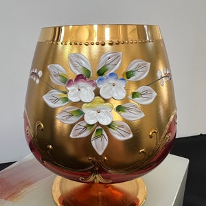 May include: A decorative, gold-colored glass vase with a wide body and a flared base. The vase is adorned with hand-painted white flowers with green, blue, and purple accents. The rim and base are trimmed with gold, and the vase sits on a clear, white surface.