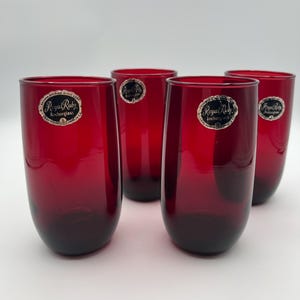 May include: A set of four cylindrical drinking glasses made of deep red translucent glass. Each glass features a circular sticker near the rim with the text Royal Ruby Anchorglass, indicating the manufacturer and the specific color collection.