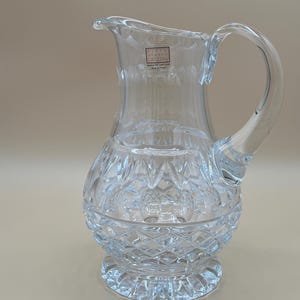 May include: Clear crystal pitcher with a rounded base and a flared spout. The pitcher features a diamond-cut pattern on the lower half and vertical lines on the upper half. A curved handle is attached to the side. The label reads "Fifth Avenue Crystal, Handcut Full Lead Crystal, Made in Poland."