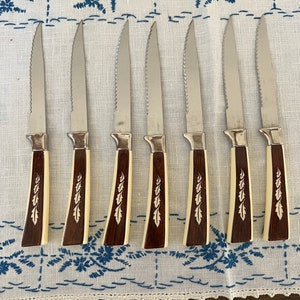 May include: Set of seven steak knives with serrated stainless steel blades. The handles are a combination of dark brown and cream-colored materials, with a leaf design. The knives are arranged on a white tablecloth with blue floral embroidery.