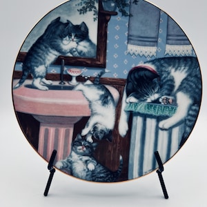 May include: A round decorative ceramic plate with a gold-trimmed edge featuring a detailed illustration of several gray and white tabby cats in a bathroom setting, including one cat looking into a mirror above a pink pedestal sink.