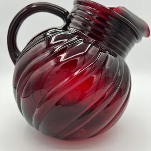 May include: A ruby red glass pitcher with a rounded body and a curved handle. The pitcher has a spiral design and a pouring spout. The glass is a deep, transparent red color. The pitcher is likely used for serving beverages.