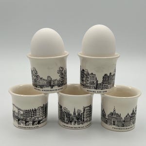 May include: A set of five off-white ceramic egg cups featuring black line-art illustrations of Amsterdam canal houses and bridges. Two cups hold white eggs. Each cup is labeled with names like Amstel-Keizersgracht, Singel 410-440, Prinseneiland, Groenburgwal, and Prinsengracht.