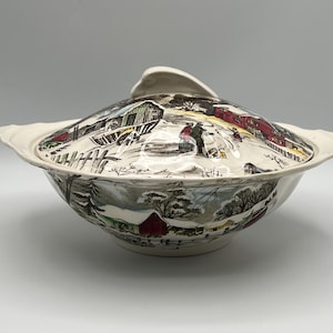 May include: A ceramic serving bowl with a matching lid, featuring a detailed winter scene with red barns, snow-covered trees, and figures in a landscape. The bowl has two side handles and a curved lid handle, decorated in a multi-colored print.