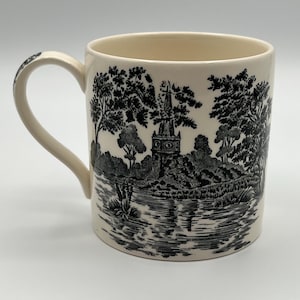 May include: Cream-colored ceramic mug with a black and white pastoral scene. The design features trees, a church steeple, and a body of water. The mug has a curved handle and a smooth, glossy finish.
