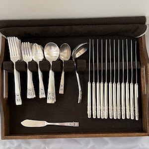 May include: A silver flatware set, including forks, spoons, knives, and a ladle, arranged in a wooden storage tray. The silverware features a decorative pattern on the handles. A butter knife is also included in the set.