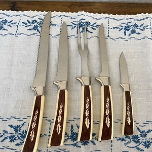 May include: A set of five stainless steel steak knives and a carving fork. The handles are cream-colored with a dark wood grain and leaf design. The knives are arranged on a white tablecloth with a blue floral pattern.