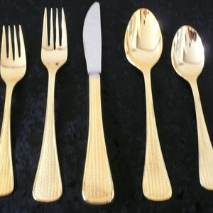 May include: A set of gold-colored flatware, including two forks, a knife, and two spoons. The handles have a textured pattern. The knife has a silver blade. The flatware is arranged on a dark surface.