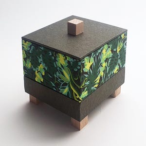 Könnte beinhalten: Eine quadratische dekorative Box mit einem dunkelgrauen Deckel und Boden sowie einer gemusterten Seite mit grünen und gelben Blättern auf schwarzem Hintergrund. Der Deckel hat einen kleinen, quadratischen Holzknopf. Die Box steht auf vier kleinen Holzfüßen.