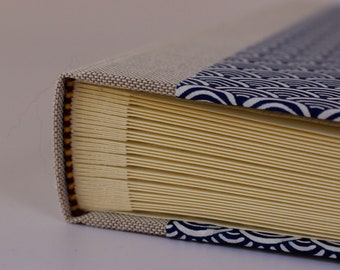 May include: Close-up of the spine of a hardcover book with a beige linen cover and a blue and white patterned fabric cover. The book is bound with a yellow and brown ribbon.