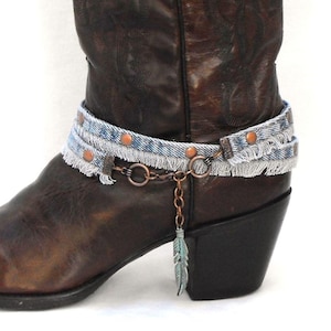 May include: A brown leather cowboy boot with a pointed toe. The boot is adorned with a denim boot cuff featuring copper studs and a dangling feather charm. The boot has a black heel and intricate stitching.