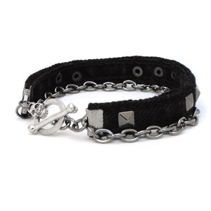 May include: Black fabric bracelet with silver-tone metal chain and square studs. The bracelet features a toggle clasp with a skull design. The bracelet is a punk rock style accessory.