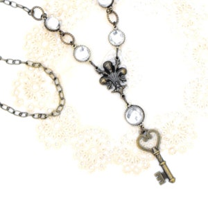 May include: Antique brass-toned necklace featuring clear, round crystals and a key pendant. The necklace has a fleur-de-lis design and a delicate chain. The crystals are set in a vintage-style setting, creating an elegant and timeless look.