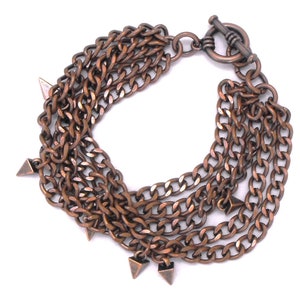 May include: A copper-colored chain bracelet with multiple strands. The bracelet features a toggle clasp and small, triangular spikes hanging from the chains. The chains are made of a metal with a rustic, aged appearance.