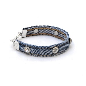 May include: A denim bracelet with silver-tone metal accents and three clear, round rhinestones. The bracelet has a silver clasp and two magnetic closures. The denim is blue with a light wash.