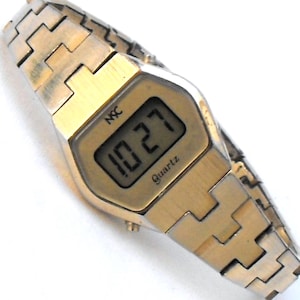 May include: A gold-tone digital wristwatch with a metal link band. The rectangular watch face features a digital display showing the time 10:27, with the text NSC printed above the display and the word Quartz printed below the digital numbers.