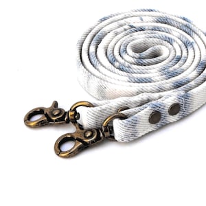 May include: A rolled-up, white and blue patterned strap with two antique brass-colored swivel clasps. The strap is made of fabric and has a denim-like appearance. The clasps are attached to the strap with metal rivets.
