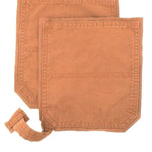 May include: Two orange-brown canvas pockets with reinforced stitching. The pockets are rectangular with a curved strap attached to one. The pockets are likely for workwear or a similar type of garment.